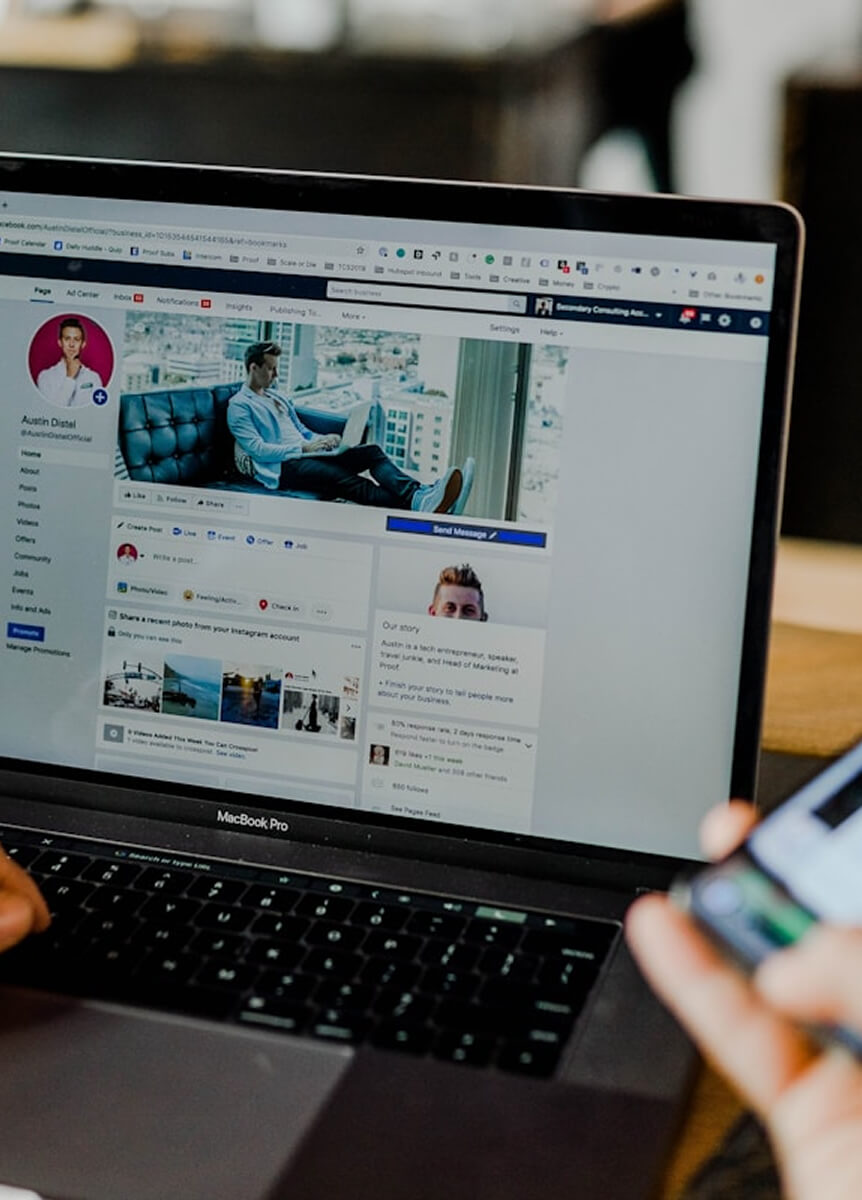 Laptop képernyőjén Facebook oldal, mellette okostelefont tartó kéz